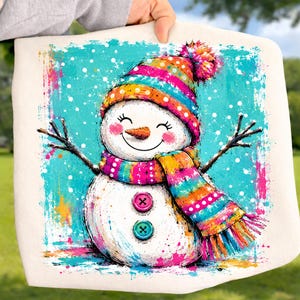 May include: A white fabric square featuring a colorful illustration of a snowman with a bright orange carrot nose, pink cheeks, and two buttons on its torso. The snowman wears a striped beanie and scarf in shades of teal, orange, and pink.