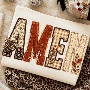 May include: A white sweatshirt featuring the word AMEN in large, decorative block letters. The letters are filled with various textures and patterns, including floral prints, brown corduroy, beige gingham, and cream cable knit fabric.