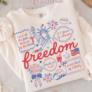 May include: A cream-colored long-sleeve sweatshirt featuring a red and blue graphic print with the Statue of Liberty, fireworks, a cowboy hat, and the text "Land of the free because of the brave," "250 Years of Freedom," "freedom," "made in USA," and "Sweet land of Liberty."