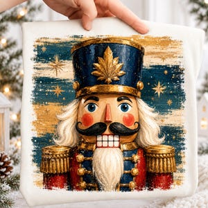 May include: A white fabric item featuring a graphic print of a nutcracker with a blue hat, gold leaf emblem, white hair, black mustache, and red uniform. The background consists of a distressed blue and gold horizontal brushstroke pattern.