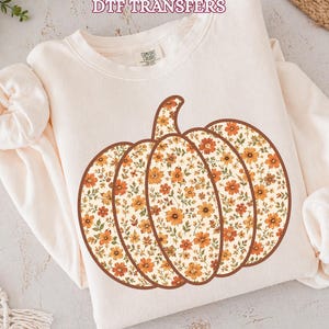 May include: A cream-colored long-sleeve sweatshirt featuring a large pumpkin graphic filled with a repeating pattern of small orange and yellow flowers with green leaves. The text DTF TRANSFERS is printed in a dark outline at the top of the image.