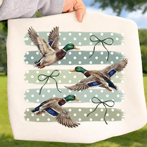 May include: A cream-colored tote bag featuring a printed design of three mallard ducks in flight. The background consists of horizontal sage green brushstroke stripes with white polka dots, accented by three thin, dark green line-art bows.