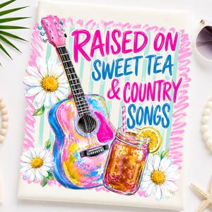 May include: A white fabric surface featuring a colorful graphic print of a pink and blue acoustic guitar, a mason jar filled with iced tea, and white daisies. The text reads RAISED ON SWEET TEA & COUNTRY SONGS in bright pink and blue lettering.