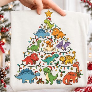 以下が含まれることがあります： 白いTシャツに、カラフルな恐竜のキャラクターたちがクリスマスツリーの形に並んだグラフィックが描かれています。恐竜たちはサンタの帽子とマフラーを身につけ、イルミネーションライト、オーナメント、プレゼントに囲まれており、頂上には黄色い星が輝いています。
