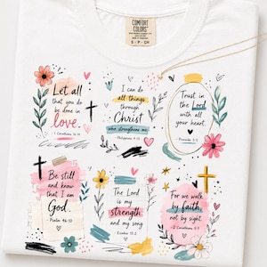 May include: A white t-shirt featuring a graphic print with six distinct sections of religious scripture verses, including Let all that you do be done in love, I can do all things through Christ, and Trust in the Lord, decorated with colorful floral accents.
