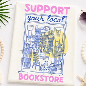 May include: A white canvas featuring a blue line drawing of a bookstore interior with shelves of books, a cat, and a basket. The text "SUPPORT" and "BOOKSTORE" is printed in pink, while "your local" is written in blue script above the shop.