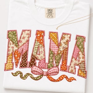 May include: A white t-shirt with the word MAMA printed in large block letters. Each letter features a patchwork design of various floral and gingham patterns in shades of green, orange, pink, and brown. A pink gingham bow sits below the text.