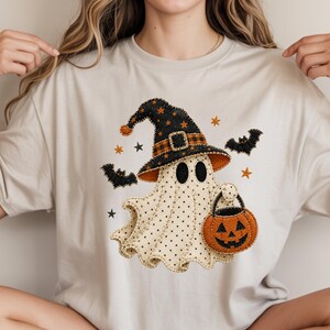 May include: A beige t-shirt featuring a graphic of a ghost wearing a black witch hat with orange stars and a plaid band. The ghost has black polka dots and carries an orange jack-o'-lantern bucket. Black bats and orange stars surround the ghost.