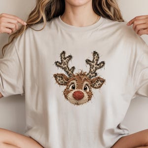 以下が含まれることがあります： トナカイの頭のグラフィックが中央に描かれたベージュの半袖Tシャツ。トナカイは赤い鼻、大きな目、ヒョウ柄の角が特徴です。デザインは、ほつれた布を縫い付けたような見た目になっています。