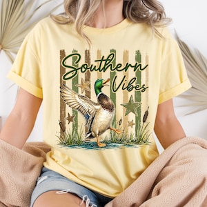 May include: Yellow t-shirt with the text "Southern Vibes" and a graphic of a duck with outstretched wings. The design includes green stars, cattails, and vertical stripes in shades of green and brown.