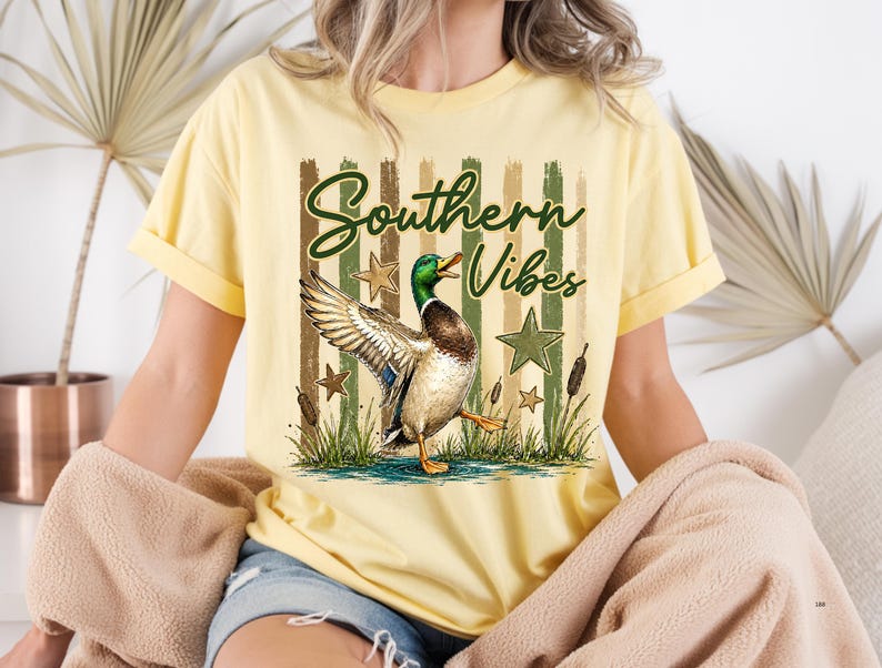May include: Yellow t-shirt with the text "Southern Vibes" and a graphic of a duck with outstretched wings. The design includes green stars, cattails, and vertical stripes in shades of green and brown.