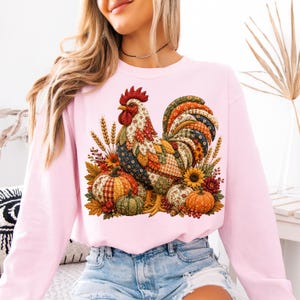 May include: A light pink long-sleeve sweatshirt featuring a central graphic of a rooster composed of various colorful fabric patterns. The rooster is surrounded by pumpkins, sunflowers, wheat stalks, and autumn leaves in shades of orange, green, and brown.