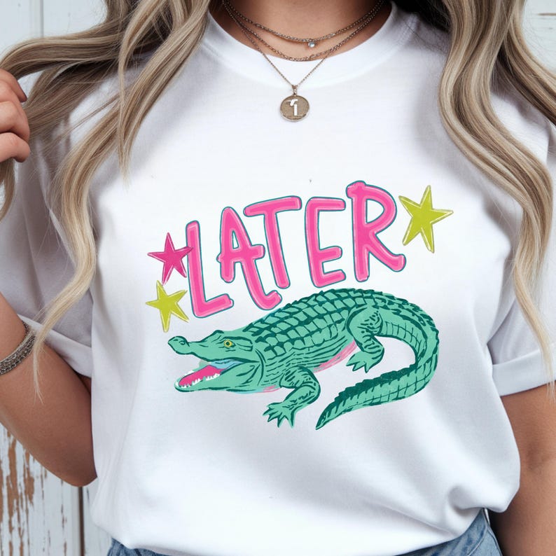 May include: White t-shirt featuring a green alligator illustration with the word "LATER" in pink, accented by yellow and pink stars. The alligator has an open mouth, and the shirt is a casual style.