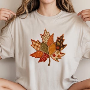 May include: A light beige short-sleeve t-shirt featuring a large central graphic of a maple leaf. The leaf is composed of various fabric-like patches in shades of orange, brown, green, and cream, with visible stitching along the edges.
