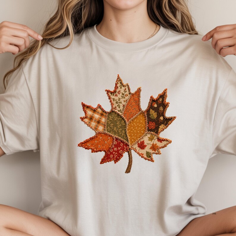 May include: A light beige short-sleeve t-shirt featuring a large central graphic of a maple leaf. The leaf is composed of various fabric-like patches in shades of orange, brown, green, and cream, with visible stitching along the edges.