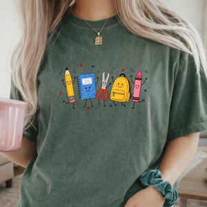 May include: A dark olive green short-sleeve t-shirt featuring a graphic print of anthropomorphic school supplies including a yellow pencil, a blue notebook, silver scissors, a yellow backpack, and a pink crayon, all with smiling faces.