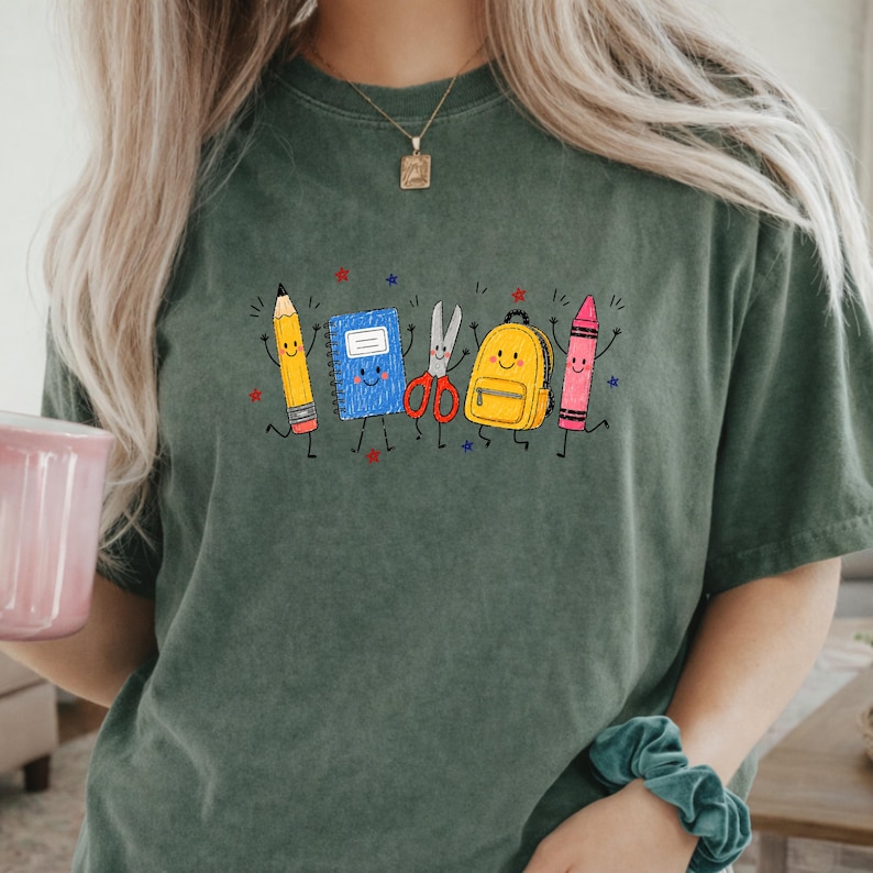 May include: A dark olive green short-sleeve t-shirt featuring a graphic print of anthropomorphic school supplies including a yellow pencil, a blue notebook, silver scissors, a yellow backpack, and a pink crayon, all with smiling faces.