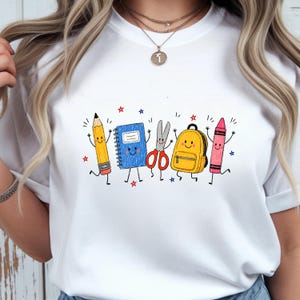 May include: A white t-shirt featuring a colorful graphic print of anthropomorphic school supplies including a yellow pencil, a blue spiral notebook, red scissors, a yellow backpack, and a pink crayon, all accented with small red and blue stars.