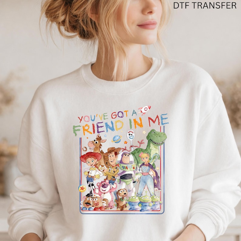 K&ouml;nnte beinhalten: Ein wei&szlig;es Sweatshirt mit Rundhalsausschnitt und einer bunten Grafik verschiedener animierter Spielzeugfiguren. &Uuml;ber den Figuren steht der Text YOU'VE GOT A FRIEND IN ME in mehrfarbigen Buchstaben. Das Design enth&auml;lt ein kleines Raketensymbol.