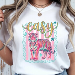 May include: White t-shirt featuring a colorful tiger illustration with the word "easy" in a decorative font. The tiger is depicted in shades of pink, blue, and green, set against a pink and white striped background with star accents.