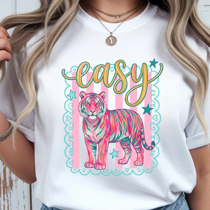 May include: White t-shirt featuring a colorful tiger illustration with the word "easy" in a decorative font. The tiger is depicted in shades of pink, blue, and green, set against a pink and white striped background with star accents.