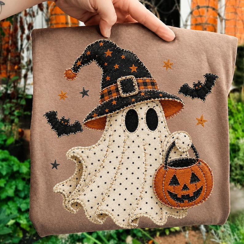 May include: A tan t-shirt featuring a central applique of a cream-colored ghost with black polka dots. The ghost wears a black witch hat with orange stars and a plaid band, holding an orange jack-o'-lantern bucket, surrounded by black bats.