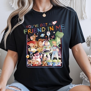 K&ouml;nnte beinhalten: Ein schwarzes Kurzarm-T-Shirt mit einer bunten Grafik verschiedener animierter Figuren aus der Toy Story-Reihe. Der Text "YOU'VE GOT A FRIEND IN ME" ist in einer mehrfarbigen, wachsmalstiftartigen Schrift &uuml;ber der Figurengruppe gedruckt.