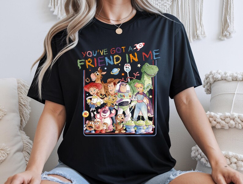K&ouml;nnte beinhalten: Ein schwarzes Kurzarm-T-Shirt mit einer bunten Grafik verschiedener animierter Figuren aus der Toy Story-Reihe. Der Text "YOU'VE GOT A FRIEND IN ME" ist in einer mehrfarbigen, wachsmalstiftartigen Schrift &uuml;ber der Figurengruppe gedruckt.
