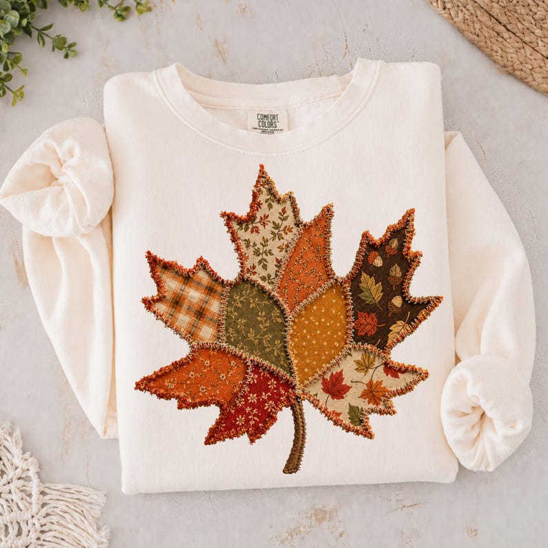 May include: A cream-colored long-sleeve sweatshirt featuring a large, central maple leaf graphic composed of various fabric patches in autumn colors including orange, brown, green, and tan with a frayed edge finish and a Comfort Colors brand label.