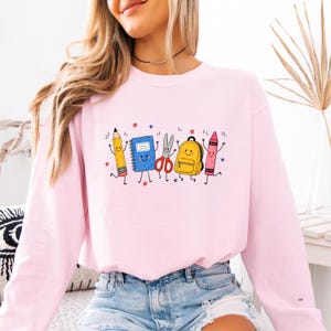 May include: A light pink long-sleeve sweatshirt featuring a graphic print of anthropomorphic school supplies including a yellow pencil, blue notebook, silver scissors, yellow backpack, and pink crayon, all with smiling faces and thin black limbs.