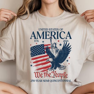 May include: Beige t-shirt with a patriotic design. The graphic includes the text "United States of America", the years 1776 and 2026, a depiction of the American flag, the Statue of Liberty, and an eagle. The phrase "We the People" is also featured.