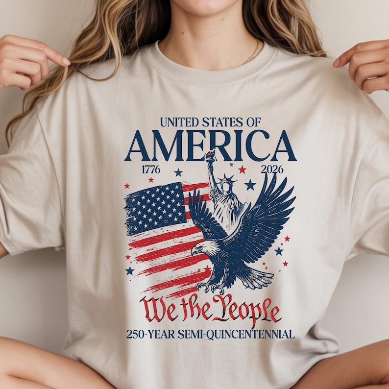 May include: Beige t-shirt with a patriotic design. The graphic includes the text "United States of America", the years 1776 and 2026, a depiction of the American flag, the Statue of Liberty, and an eagle. The phrase "We the People" is also featured.
