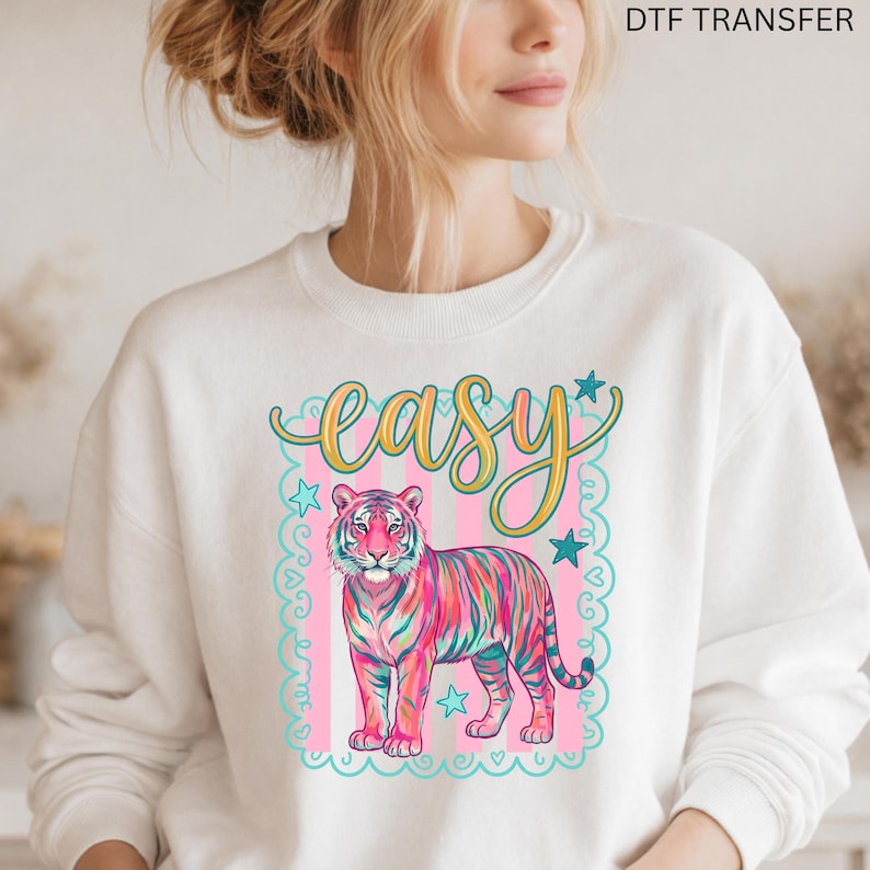 May include: White sweatshirt featuring a colorful tiger design with the word "easy" in a script font. The tiger has pink, blue, and green stripes. The design is framed by a pink and teal border with stars.