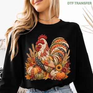 May include: A black long-sleeve sweatshirt featuring a colorful graphic of a rooster with a patchwork-style pattern. The rooster is surrounded by autumn-themed elements including wheat stalks, orange and green pumpkins, and sunflowers.
