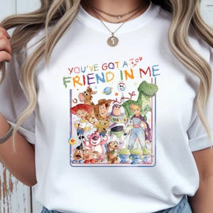 K&ouml;nnte beinhalten: Ein wei&szlig;es Kurzarm-T-Shirt mit einer bunten Aquarell-Grafik verschiedener animierter Spielzeugfiguren. &Uuml;ber den Figuren steht der Text YOU'VE GOT A FRIEND IN ME in bunten Buchstaben, erg&auml;nzt durch ein kleines Raketensymbol.