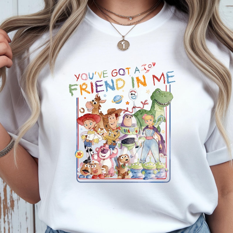 K&ouml;nnte beinhalten: Ein wei&szlig;es Kurzarm-T-Shirt mit einer bunten Aquarell-Grafik verschiedener animierter Spielzeugfiguren. &Uuml;ber den Figuren steht der Text YOU'VE GOT A FRIEND IN ME in bunten Buchstaben, erg&auml;nzt durch ein kleines Raketensymbol.