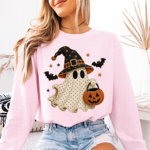May include: A light pink long-sleeve sweatshirt featuring a central graphic of a cream-colored ghost with black polka dots. The ghost wears a black witch hat with orange plaid trim and holds an orange jack-o'-lantern bucket, surrounded by bats.