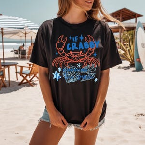 Puede incluir: Camiseta negra con un dise&ntilde;o de cangrejo y conchas marinas. El dise&ntilde;o presenta un cangrejo rojo, texto azul que dice "If Crabby" y conchas marinas azules. La camiseta se usa en un d&iacute;a soleado de playa.