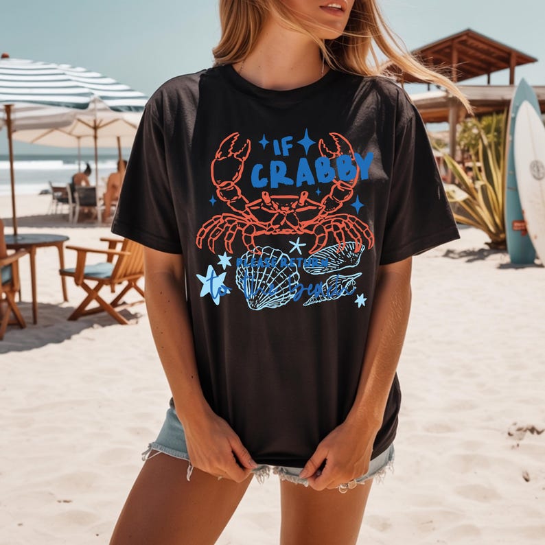Puede incluir: Camiseta negra con un dise&ntilde;o de cangrejo y conchas marinas. El dise&ntilde;o presenta un cangrejo rojo, texto azul que dice "If Crabby" y conchas marinas azules. La camiseta se usa en un d&iacute;a soleado de playa.