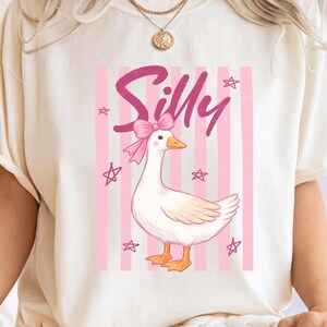 May include: Cream-colored t-shirt featuring a whimsical illustration. The design includes a cartoon goose with a pink bow, set against a backdrop of pink stripes and stars. The word "Silly" is written in pink script above the goose.