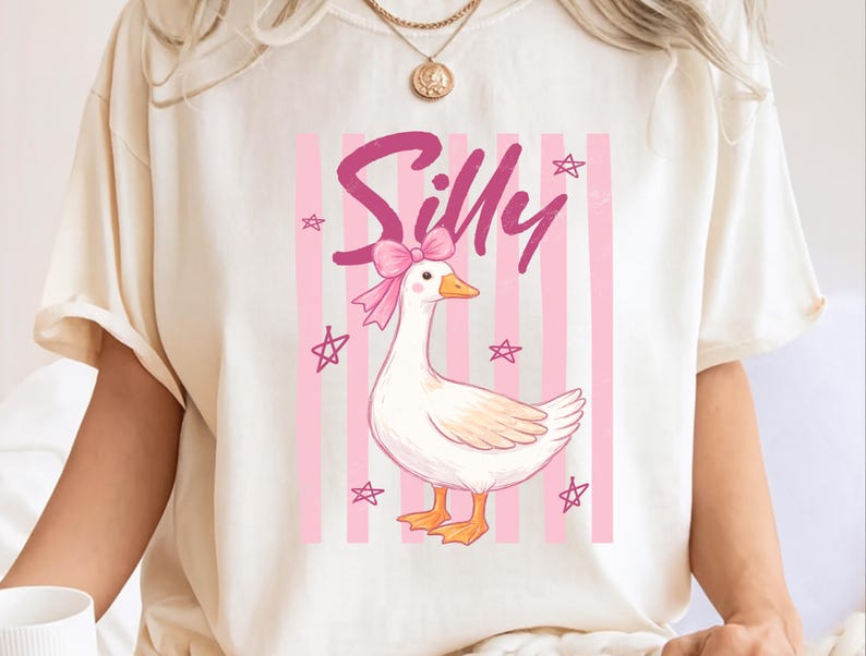 May include: Cream-colored t-shirt featuring a whimsical illustration. The design includes a cartoon goose with a pink bow, set against a backdrop of pink stripes and stars. The word "Silly" is written in pink script above the goose.