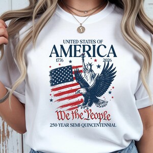 May include: White t-shirt featuring a patriotic design. The design includes the text "UNITED STATES OF AMERICA" above a depiction of the American flag, the Statue of Liberty, and an eagle. The text "We the People" and "250-YEAR SEMI-QUINCENTENNIAL" are also included.