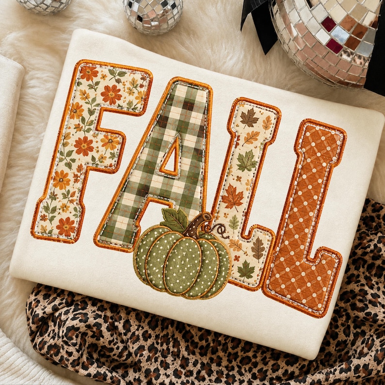 May include: A cream-coloured sweatshirt featuring the word FALL appliqued in large block letters. Each letter has a unique pattern: floral, green plaid, autumn leaves, and orange polka dots, with a green polka-dot pumpkin at the base.
