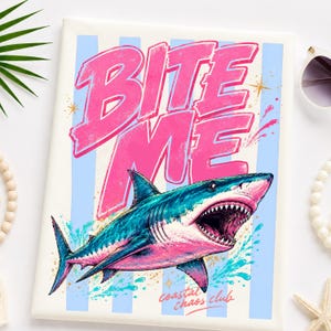 May include: A white t-shirt with a graphic of a shark with an open mouth, teal and pink coloring, and the words "BITE ME" in pink. The shirt also has the words "coastal chaos club" at the bottom.