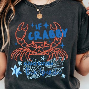Puede incluir: Camiseta gris oscuro con un dise&ntilde;o de cangrejo rojo y el texto "IF CRABBY PLEASE RETURN TO BEACH". El dise&ntilde;o incluye detalles en azul y gr&aacute;ficos de conchas marinas. Una camiseta gr&aacute;fica informal con tem&aacute;tica de playa.