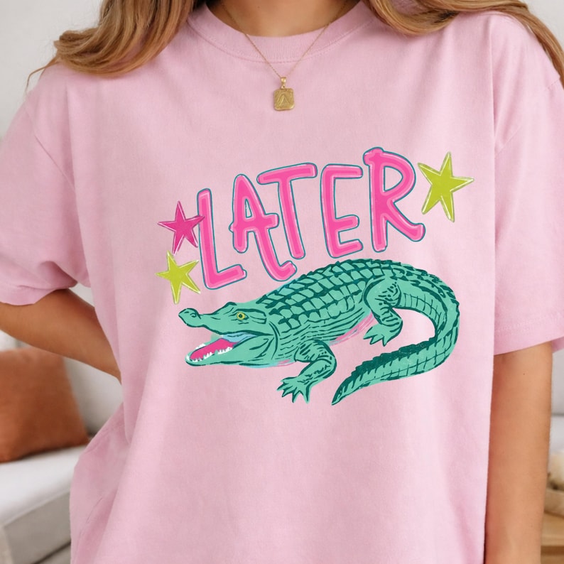 May include: Pink t-shirt featuring a teal alligator graphic with the word "LATER" in pink, outlined in white. The word "LATER" has a star on each side, one pink and one yellow.
