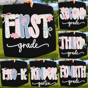 May include: Black fabric sign with the words "FIRST grade" in white script. The letters are decorated with floral and patterned designs in pink, blue, and white, with bows and flowers. The sign is held up against a green background.