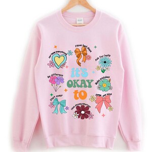 May include: A light pink sweatshirt with the words "it's OKAY to" and various phrases and illustrations. The design includes a heart, flowers, bows, and the phrases "Have bad days", "Ask for help", and "Make mistakes".