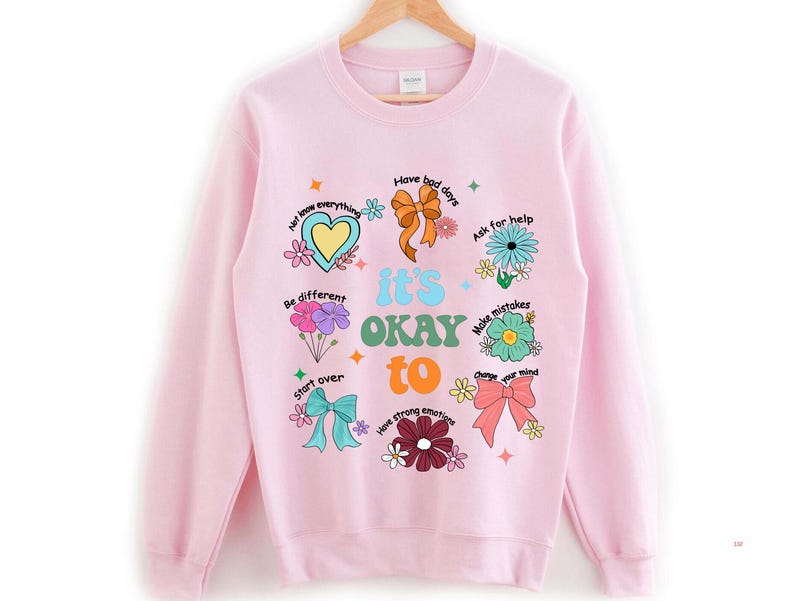 May include: A light pink sweatshirt with the words "it's OKAY to" and various phrases and illustrations. The design includes a heart, flowers, bows, and the phrases "Have bad days", "Ask for help", and "Make mistakes".