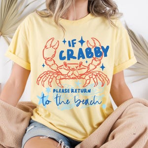 Puede incluir: Camiseta amarilla clara con un dise&ntilde;o de cangrejo rojo y azul. El texto dice "If Crabby Please Return to the Beach". La camiseta tiene mangas enrolladas y un corte holgado.